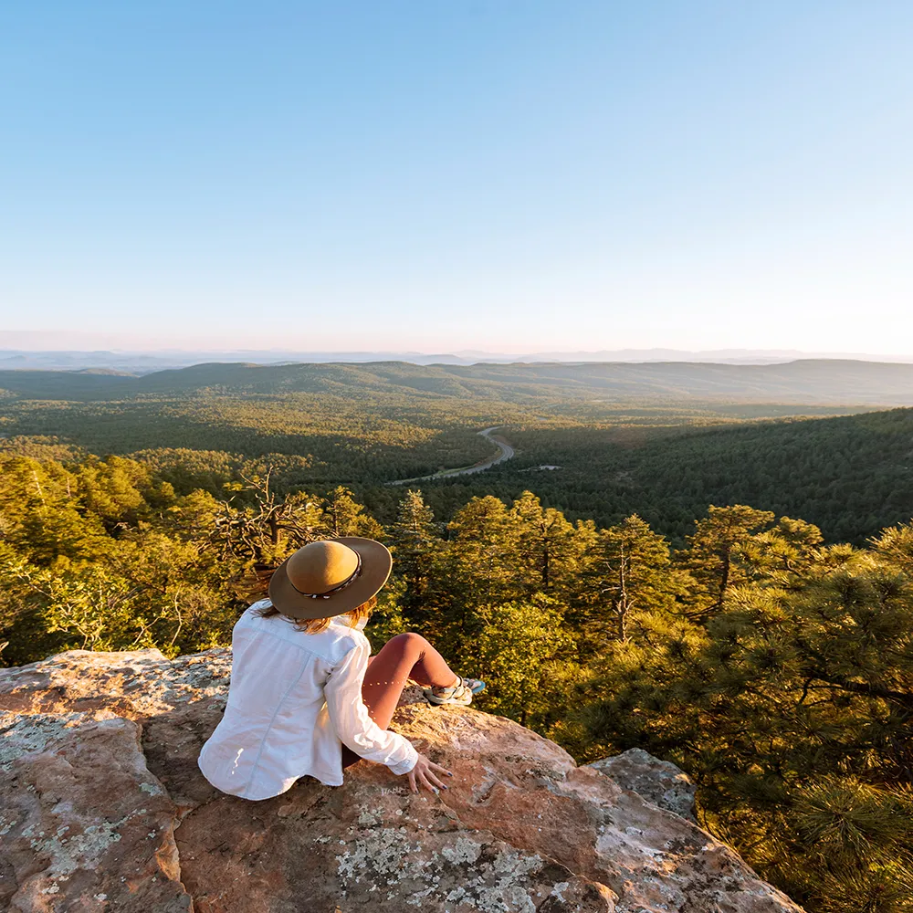 woman with hat sits on cliff edge looking out at mountain range